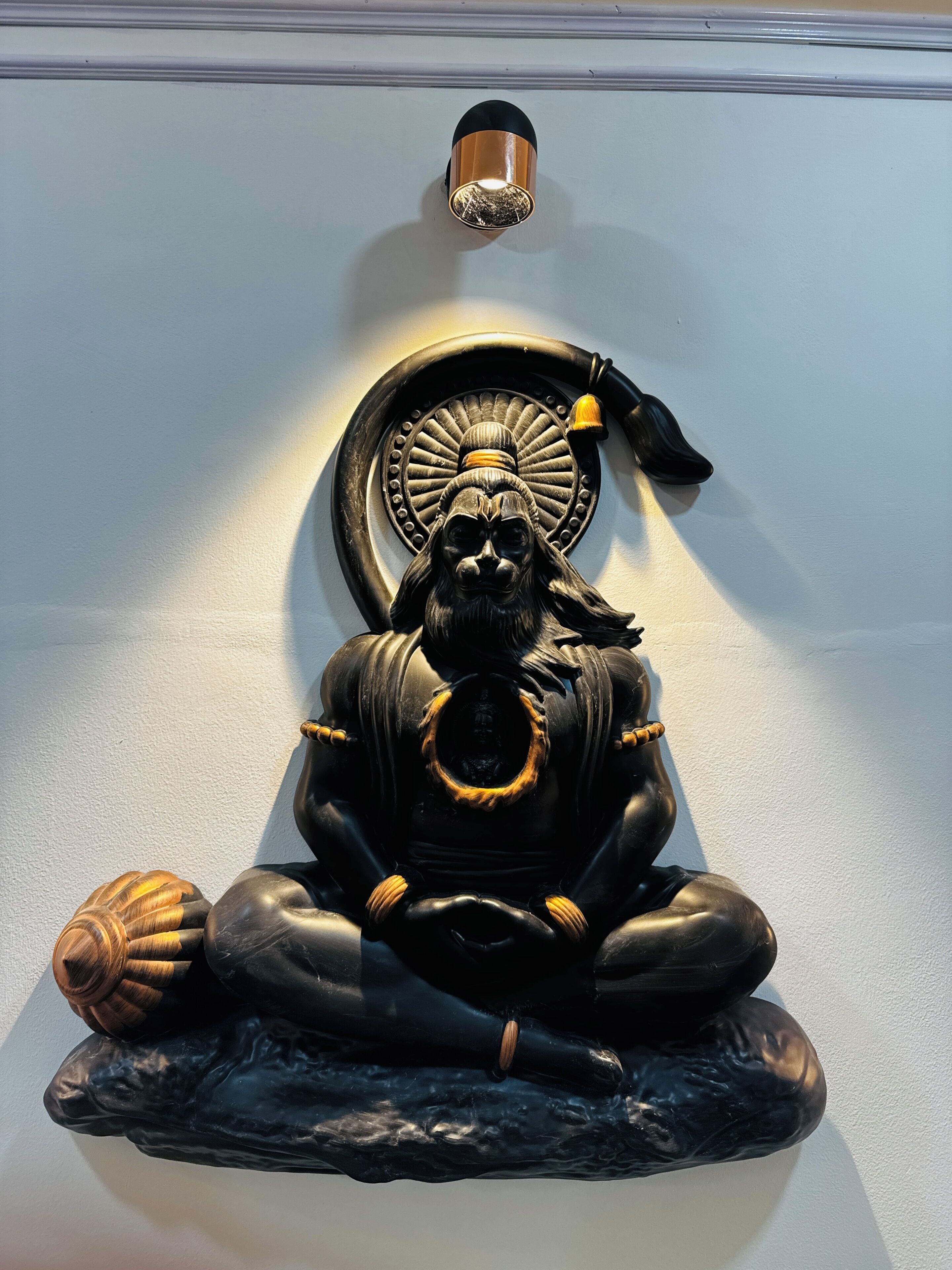 En statue af en siddende figur i meditationsstilling, med en cirkelformet genstand bagved og en lampe over sig.