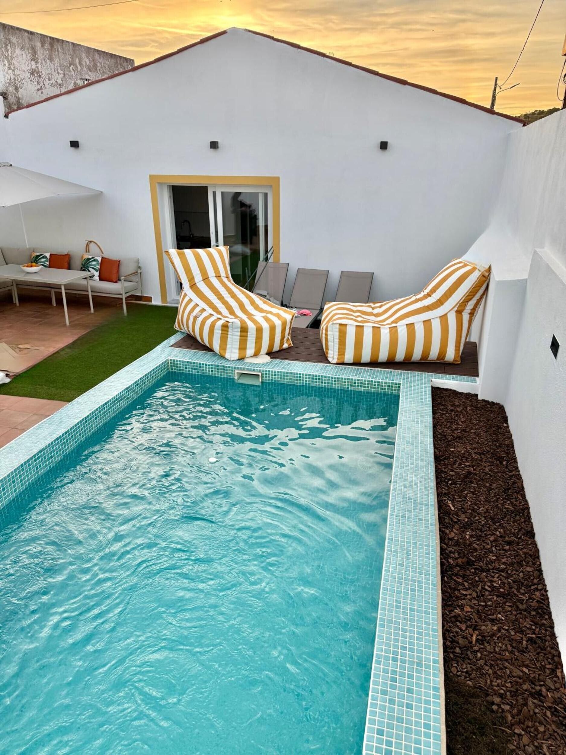Una piscina con sillas y cojines de sol, un patio con mesa y sillas, y vistas al cielo.
