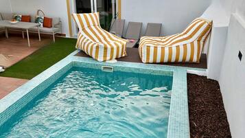 Una piscina con sillas y cojines de sol, un patio con mesa y sillas, y vistas al cielo.