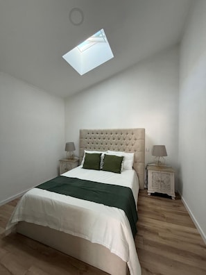 Quarto com cama, claraboia, duas mesinhas de cabeceira e um criado-mudo.
