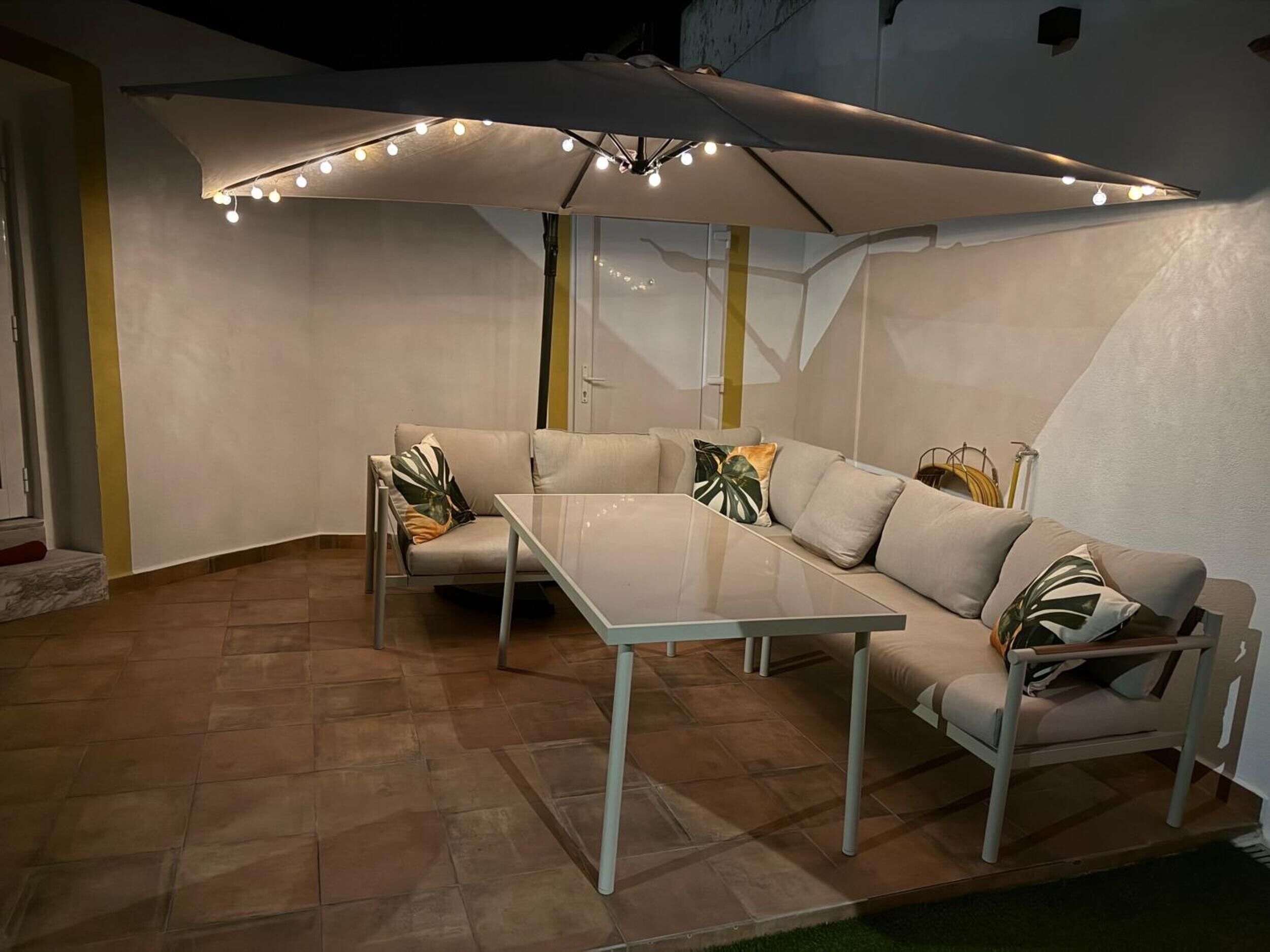 A modern outdoor seating area with a glass table and cushioned sofas, under a large umbrella with lights.