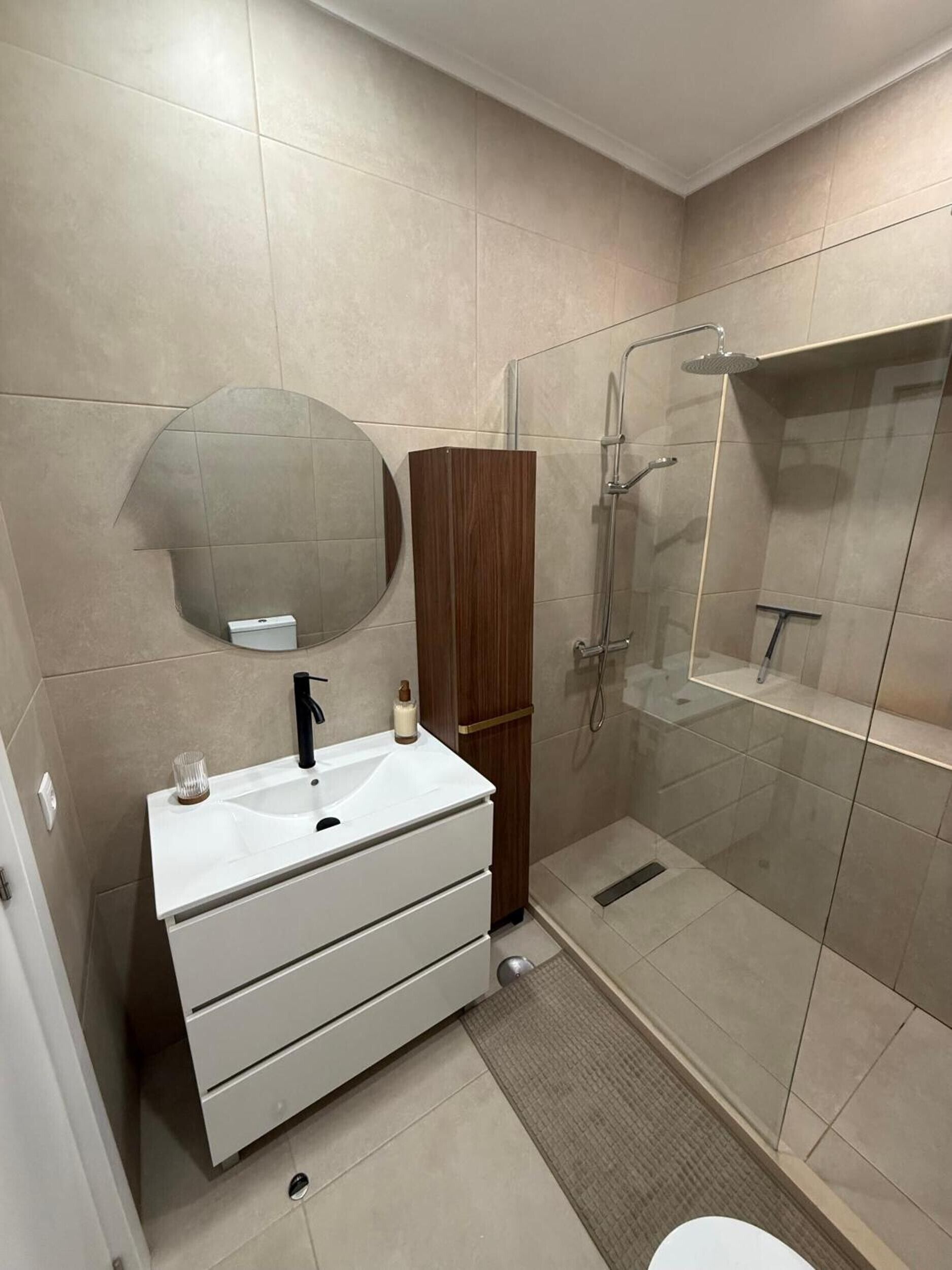 Ein modernes Badezimmer mit einem weißen Waschtisch, einem Spiegel, einem Holzschrank, einer Duschkabine aus Glas und einer Toilette.