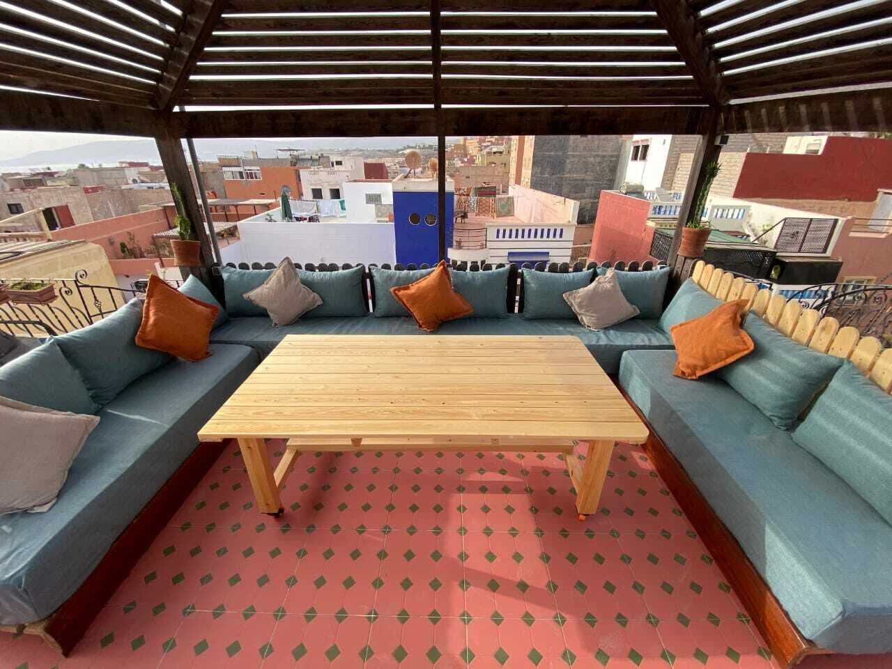 Un área de estar al aire libre techada con mesa de madera y sofás acolchados, con vista a un paisaje urbano.
