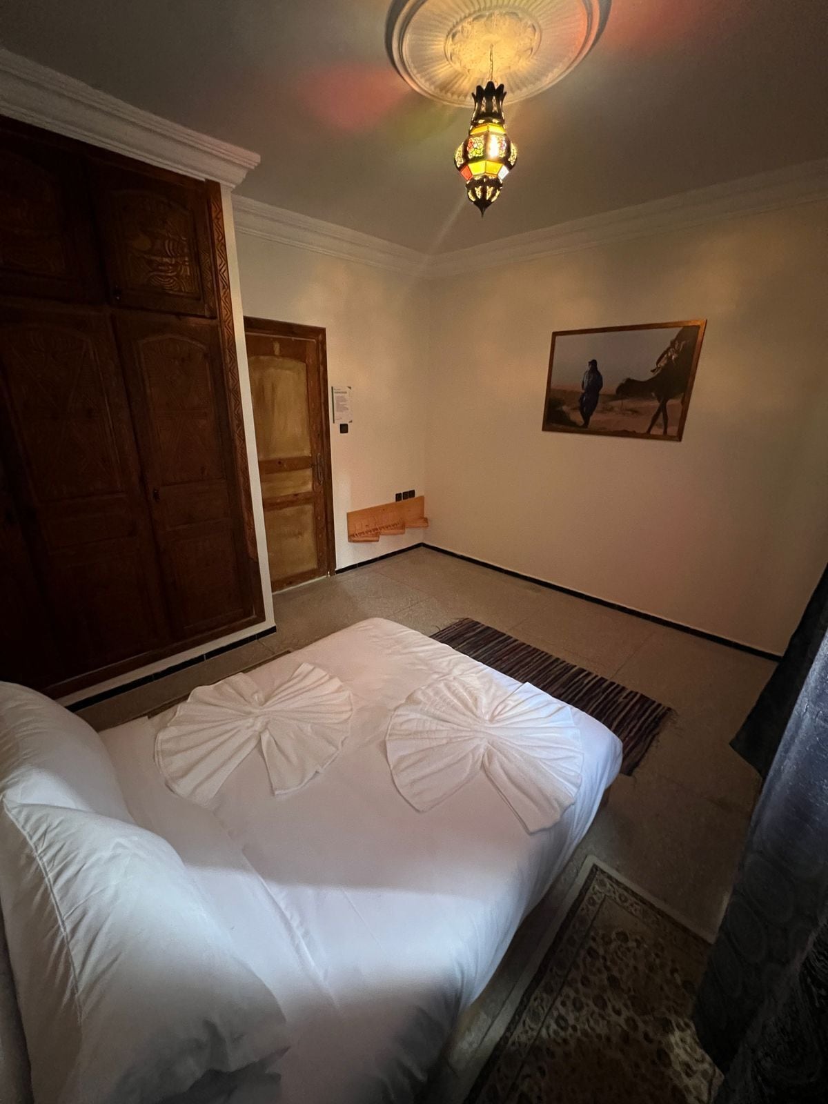 A hotel room with a bed, a wooden wardrobe, a painting on the wall, and a ceiling light fixture.
