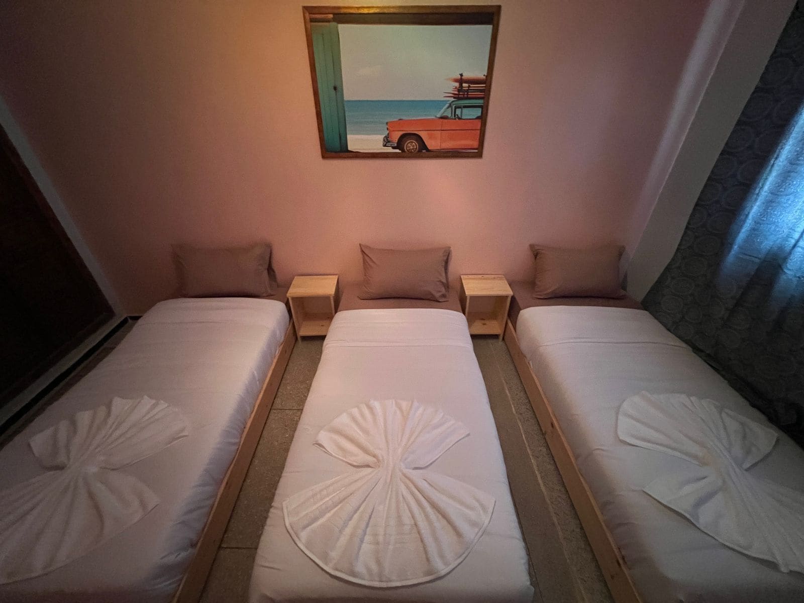 A hotel room with two single beds, each with a pillow and a wooden nightstand. A painting of a beach scene is above the beds.