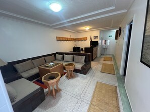 A modern living room with a sectional sofa, a coffee table, and a small side table. There is a bar area with a television and a sink in the background.