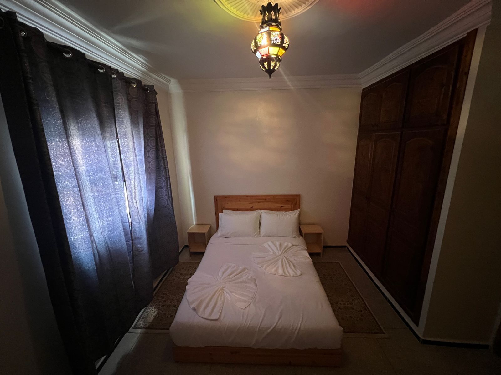 A hotel room with a bed, curtains, a nightstand, and a wall-mounted lamp.