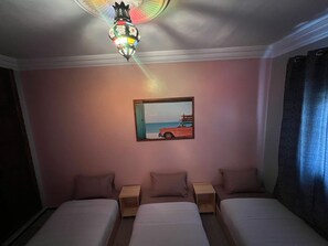 A hotel room with three single beds, a ceiling light fixture, a painting of a beach scene, and a window with curtains.