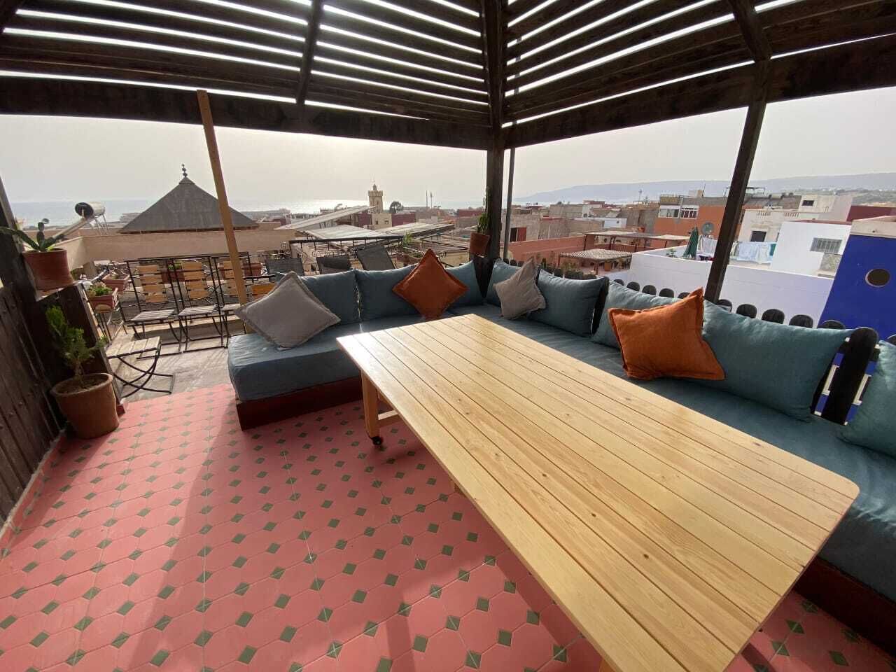 Un área de estar al aire libre con mesa de madera y sofás con cojines azules, una planta en maceta y vistas de la ciudad.