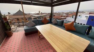 Un área de estar al aire libre con mesa de madera y sofás con cojines azules, una planta en maceta y vistas de la ciudad.