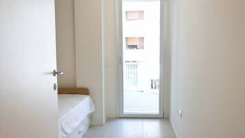 Ein schmales Entree mit einer Tür, die auf einen Balkon führt. Das Zimmer verfügt über ein weißes Bett und eine kleine Bank.