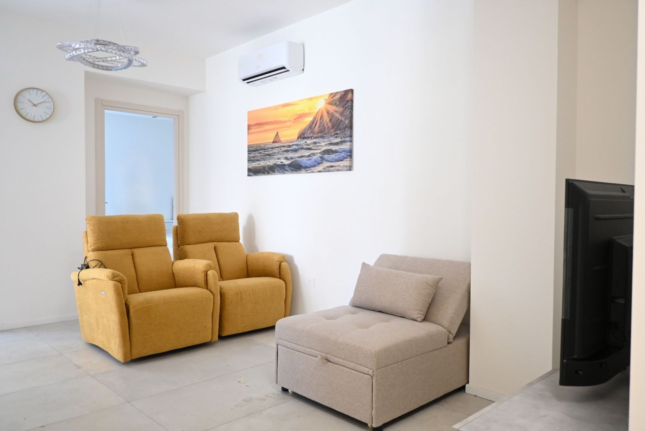 A living room with a sofa, a recliner, a flat-screen TV, a painting of a sunset, and a clock on the wall.