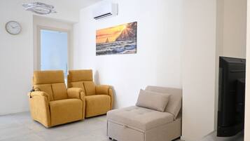 A living room with a sofa, a recliner, a flat-screen TV, a painting of a sunset, and a clock on the wall.