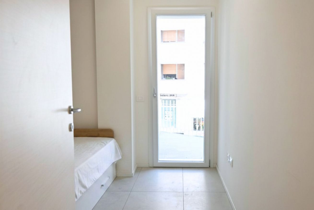 A narrow hallway with a door leading to a balcony. The room has a white bed and a small bench.