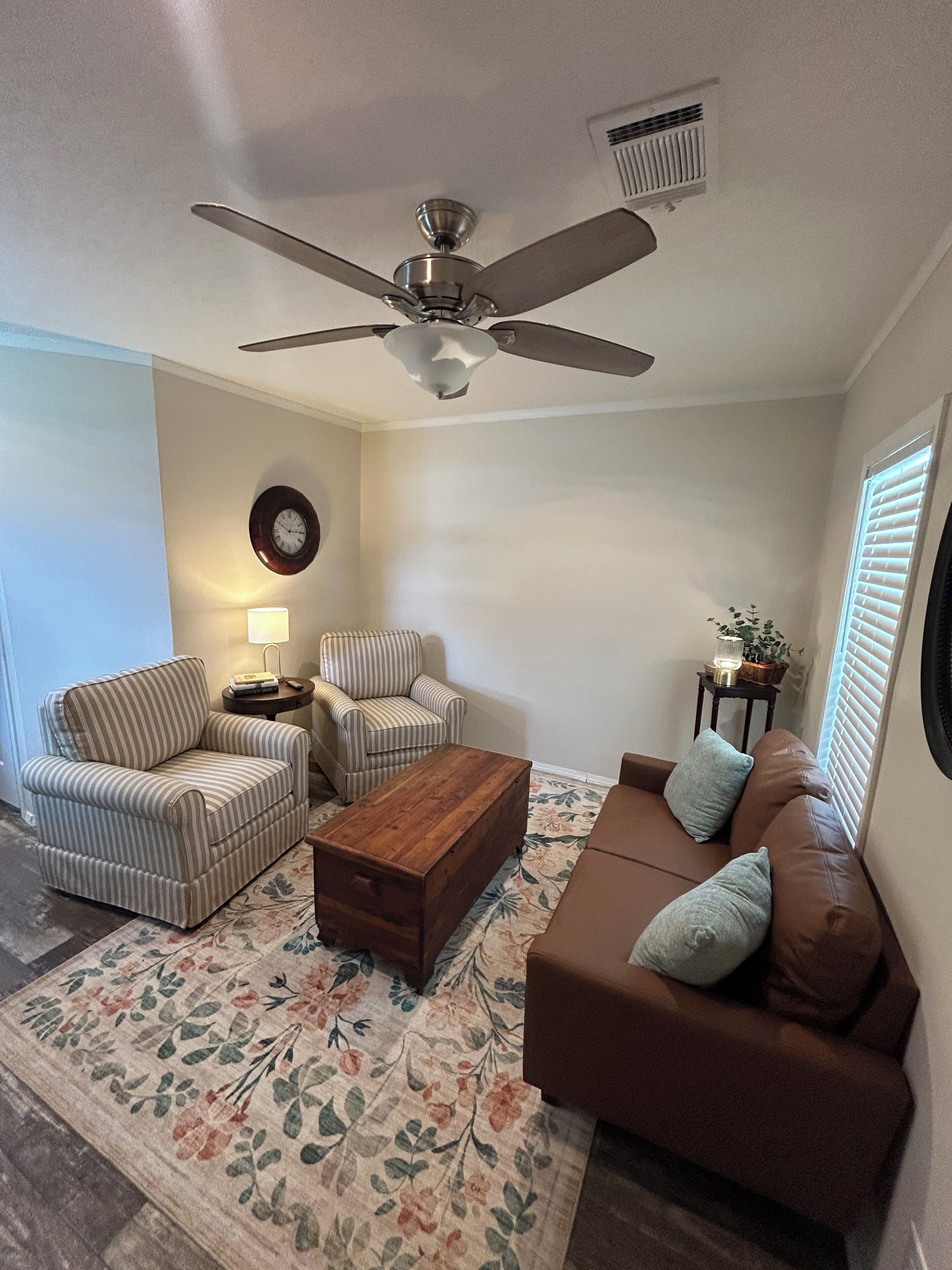 Uma sala de estar com ventilador de teto, duas poltronas listradas, um sofá marrom com almofadas, uma mesa de centro de madeira, um tapete floral, uma mesinha de cabeceira com abajur e um relógio na parede.