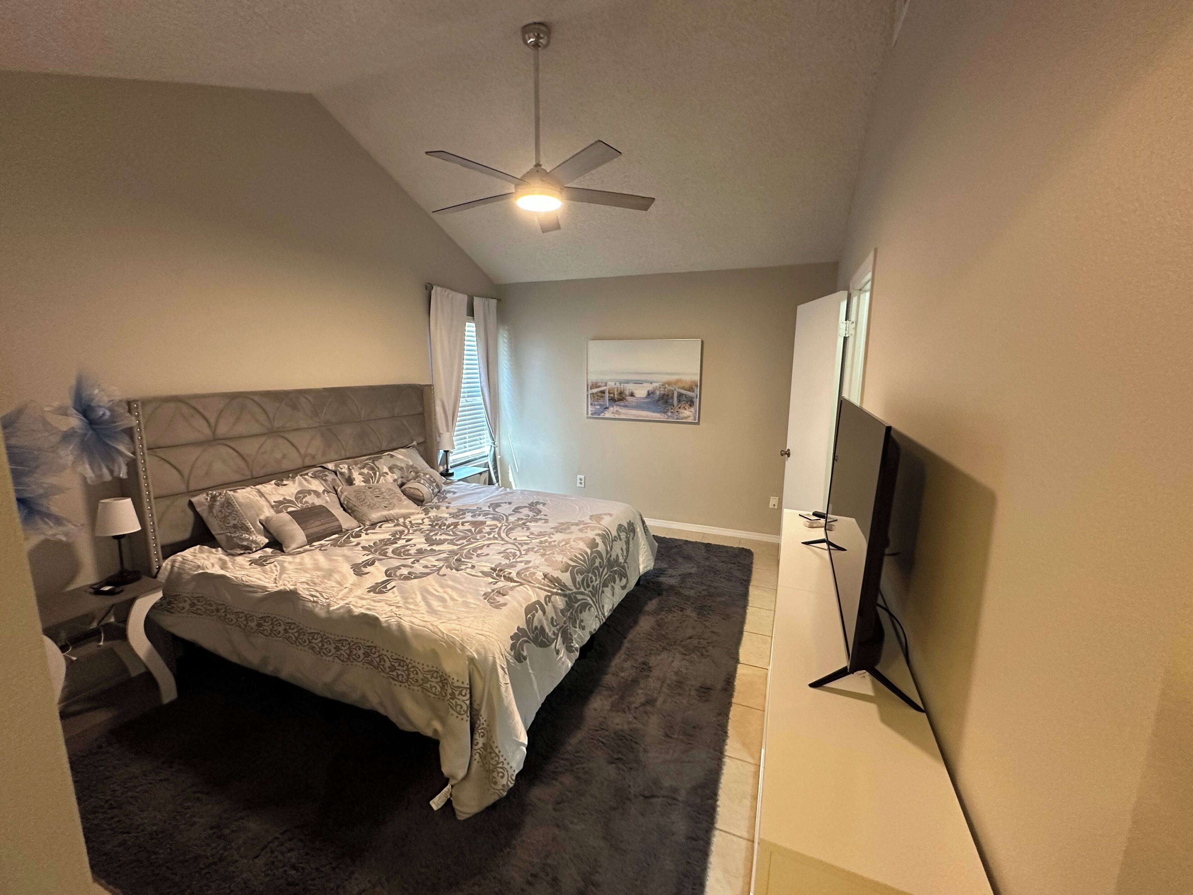 A bedroom with a bed, a ceiling fan, a flat-screen TV, and a painting on the wall.