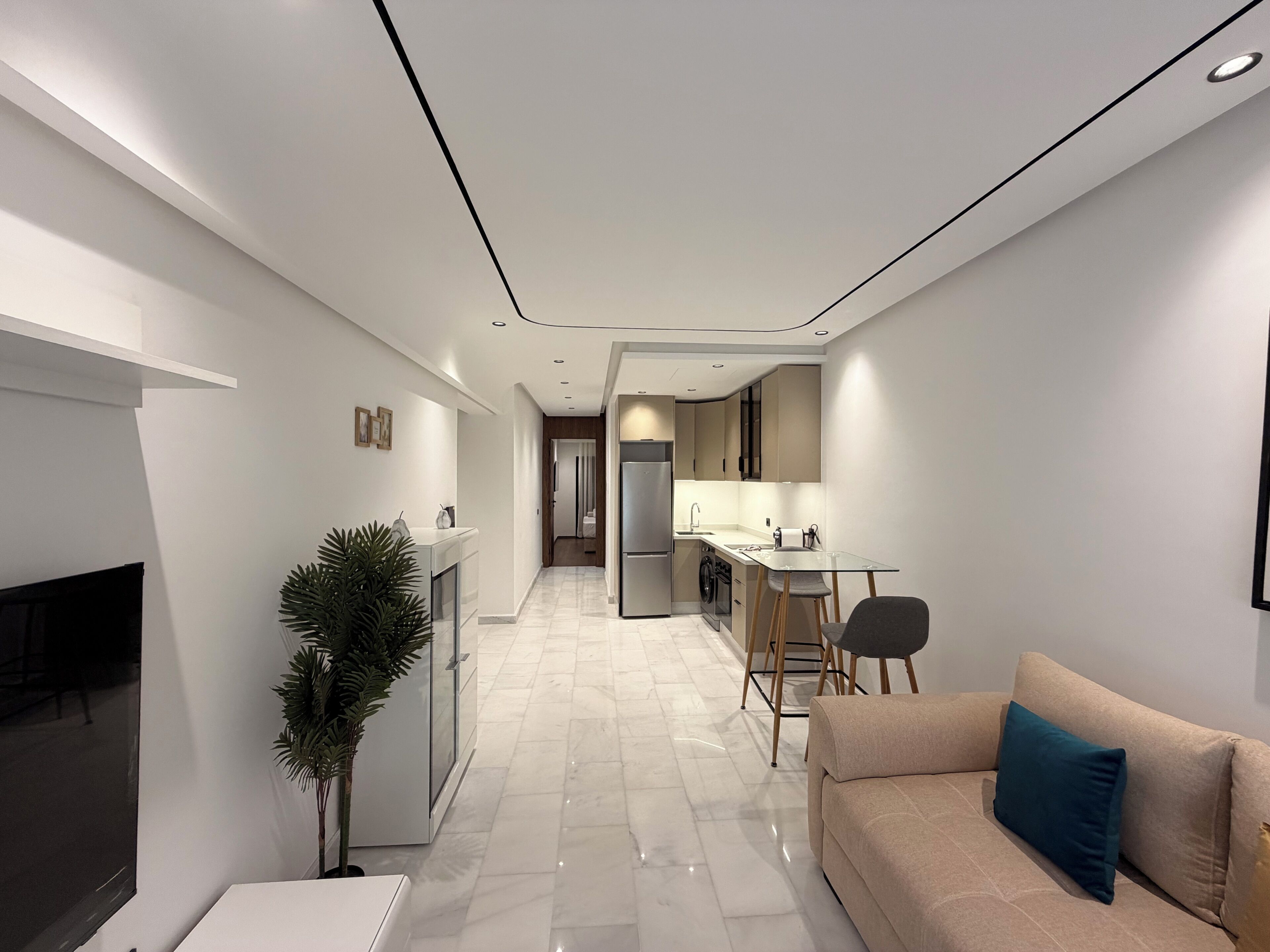 A modern living room with a sofa, a coffee table, and a TV. There is a kitchen area with a refrigerator, stove, and sink. The floor is tiled, and there is a plant in the corner.