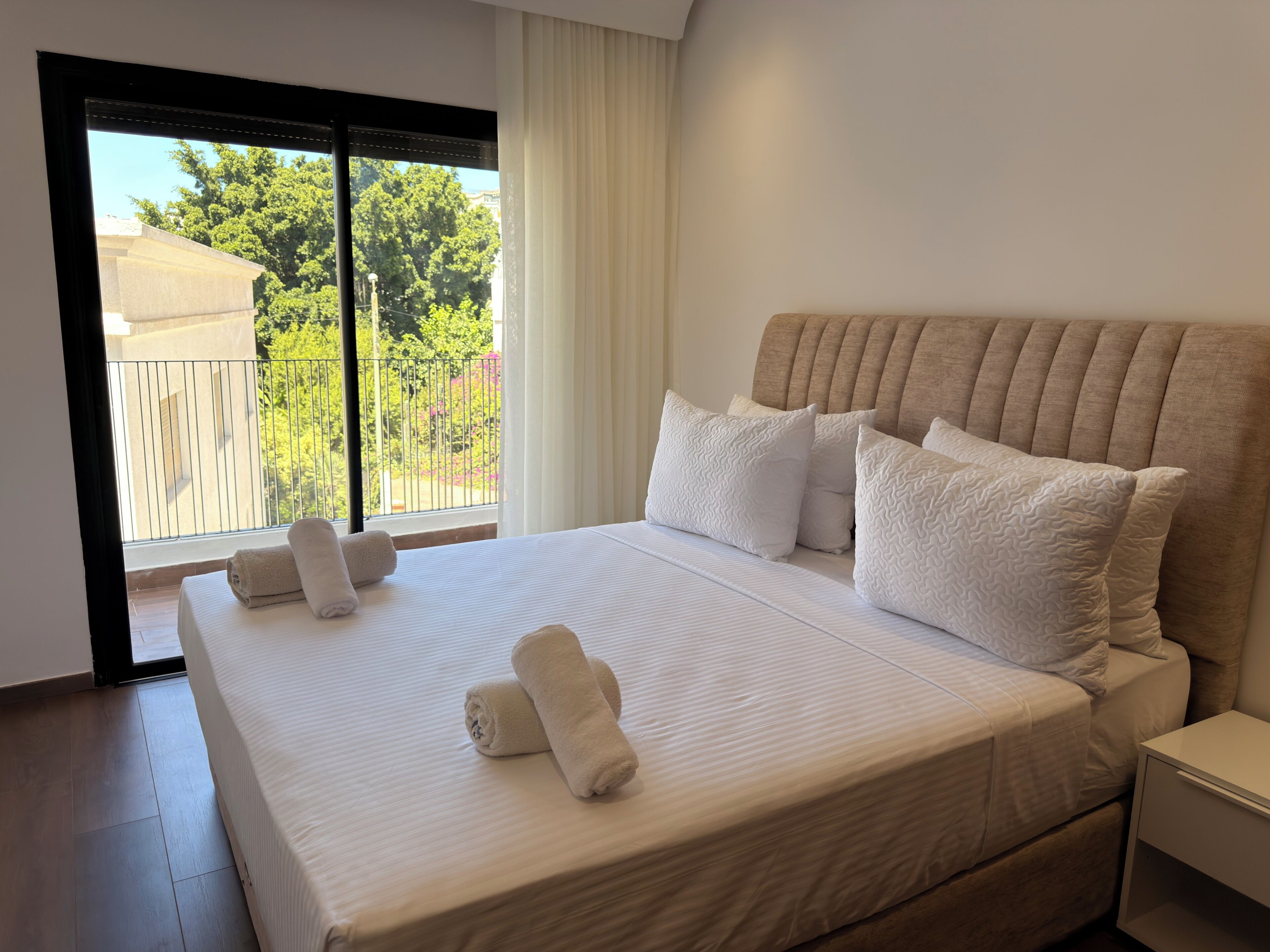 A hotel room with a large bed, a nightstand, a window with a view of greenery and buildings, and a door leading to an outdoor area.