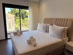 A hotel room with a large bed, a nightstand, a window with a view of greenery and buildings, and a door leading to an outdoor area. - Golden Maarif Palmiers Apartment (Casablanca)