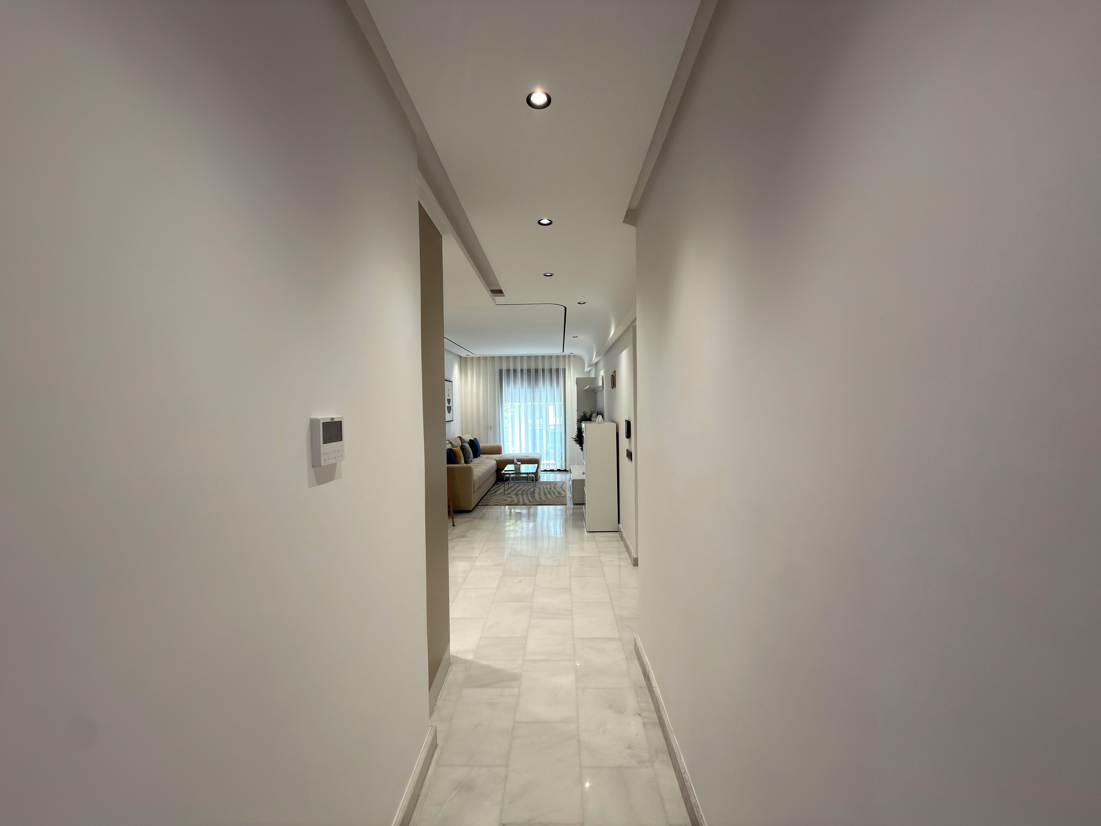 A modern hotel corridor with a sofa set, a refrigerator, and a dining area visible in the distance.