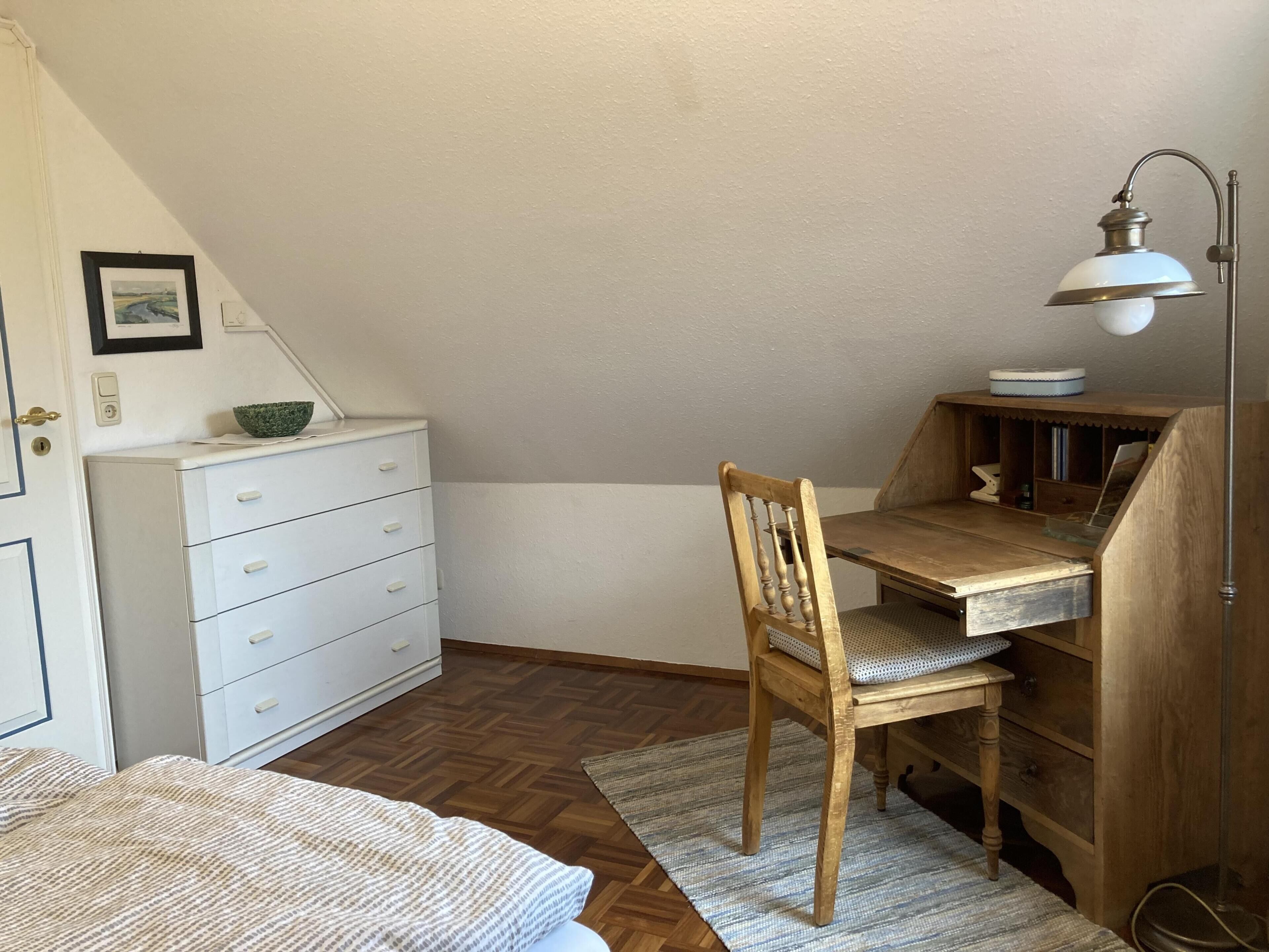 A room with a wooden desk, a chair, a white dresser, and a lamp.
