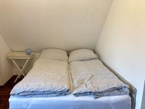 Ein kleines Zimmer mit einem Einzelbett, einer weißen Wand und einem Holzboden. Ein kleiner weißer Nachttisch mit einem blauen Gegenstand darauf.