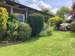 Um jardim bem cuidado com arbustos e flores variadas, uma casa de tijolos e uma estrutura de pérgola.