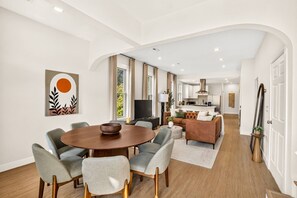 A modern living room with a round dining table, grey upholstered chairs, a sofa, and a kitchen in the background.