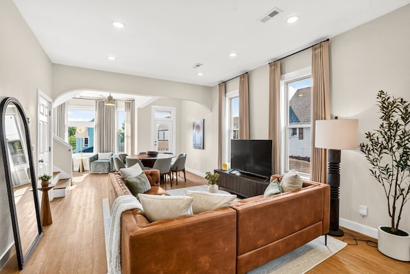A spacious living room with a brown leather sofa, a flat-screen TV, and a dining area with a table and chairs.