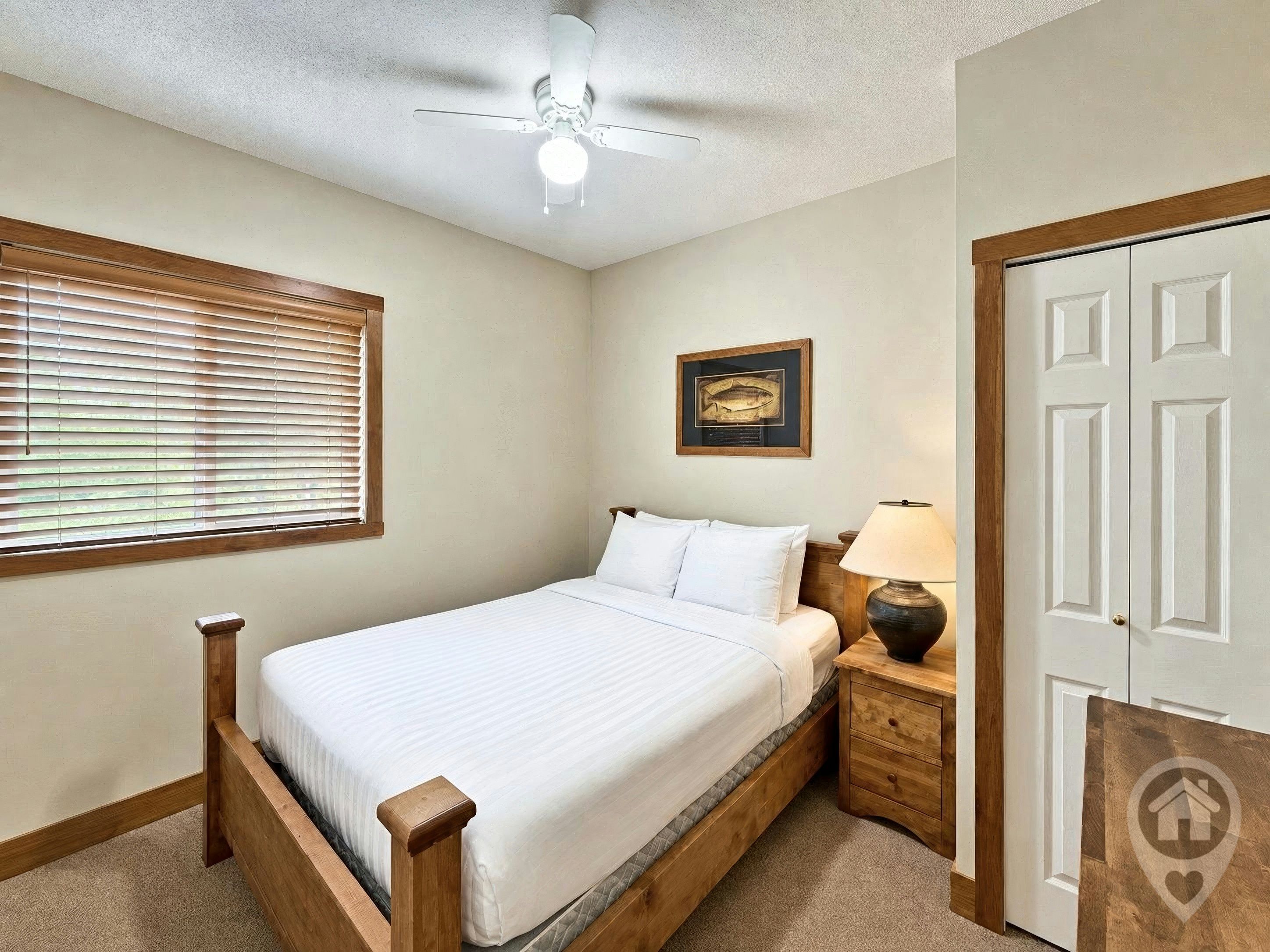 Une chambre à coucher comprenant un lit, une table de chevet, une lampe, un ventilateur de plafond, une fenêtre avec des stores et une porte.