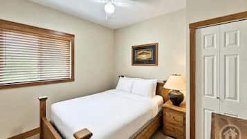 Une chambre à coucher comprenant un lit, une table de chevet, une lampe, un ventilateur de plafond, une fenêtre avec des stores et une porte.