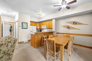 A compact kitchen with wooden cabinets, a central island, and a dining area with a wooden table and chairs. - Ski-In Condo w/ Private Hot Tub - Kimberley Ridge (Kimberley)