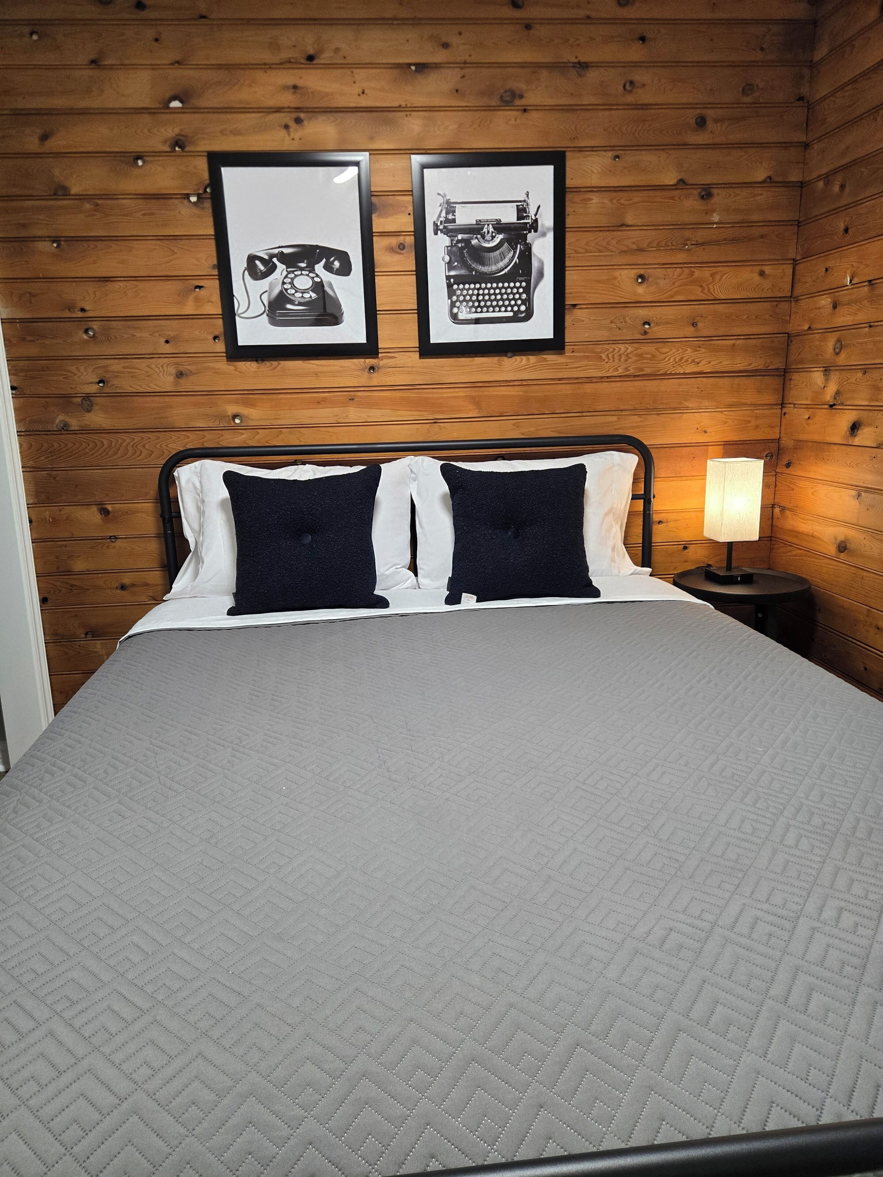 A bed with a quilted grey cover, two black pillows, and a nightstand with a lamp.