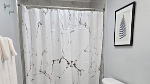 Un bagno con il water, una tenda da doccia con motivo marmorizzato, un quadro con foglie appeso al muro e un portasciugamani con asciugamani.
