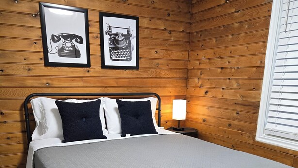A bedroom with a bed, wooden panel walls, two framed pictures, a bedside lamp, and a window with blinds.