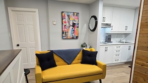 Un divano giallo con cuscini neri in una stanza con una porta bianca, un quadro astratto dai colori vivaci e un angolo cottura con mobili bianchi.