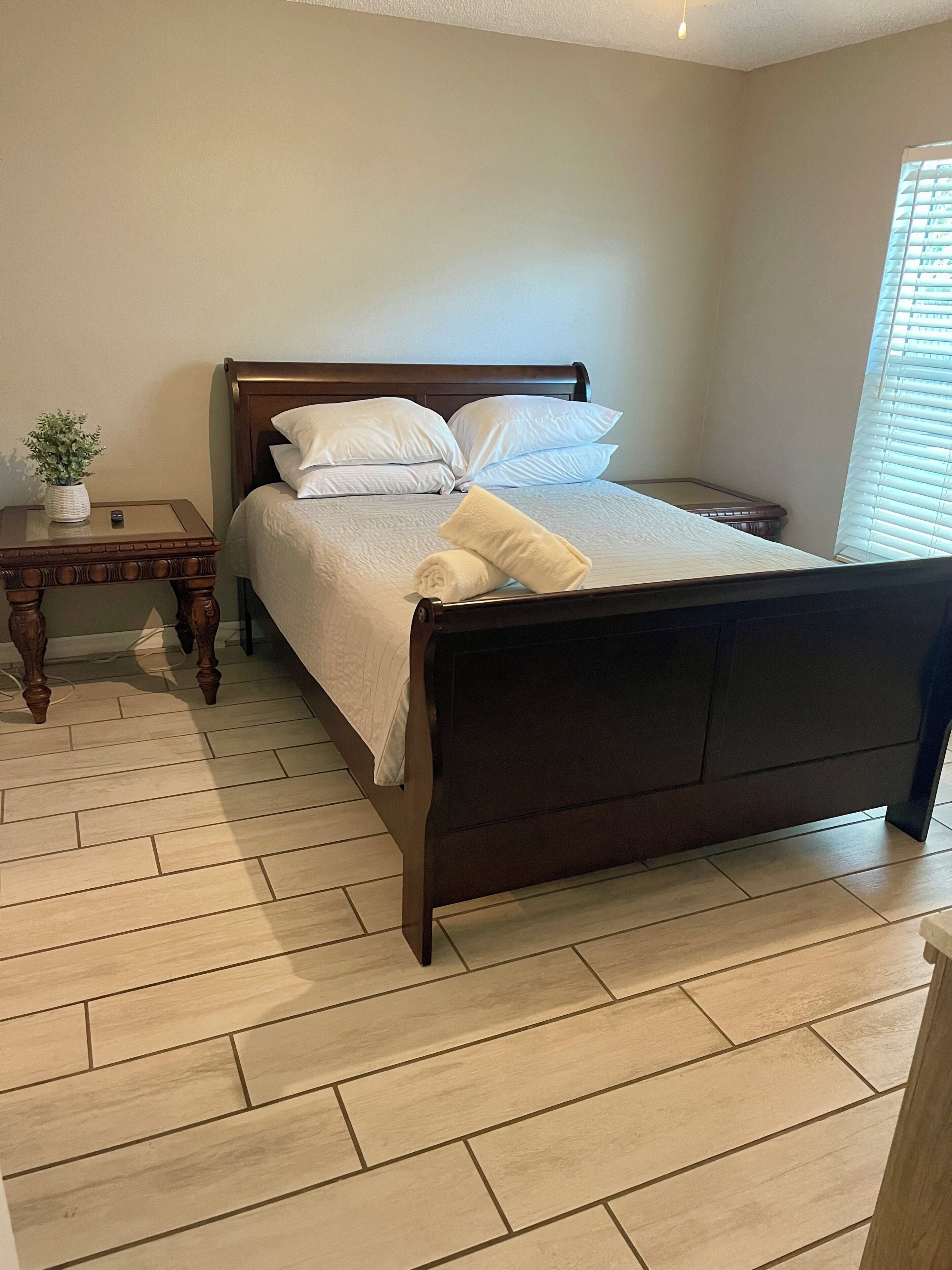 A neatly made bed with white linens and pillows, a small wooden nightstand with a plant, and a light-colored herringbone-patterned floor.