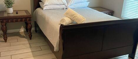 A neatly made bed with white linens and pillows, a small wooden nightstand with a plant, and a light-colored herringbone-patterned floor.