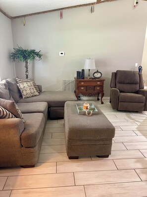 A living room with a sectional sofa, an ottoman, a side table with a lamp, a clock, and a small plant. - Coastal escape near New Smyrna Beach (Edgewater)