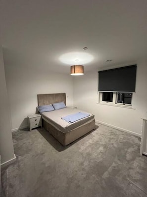A bedroom with a bed, bedside table, and a window with a blind. - Cosy Barking Apartment With City Views (Barking)