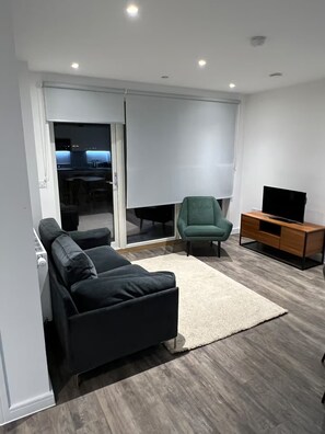 A modern living room with a sofa, a green armchair, a TV on a wooden stand, and a sliding glass door leading to a dining area. - Cosy Barking Apartment With City Views (Barking)