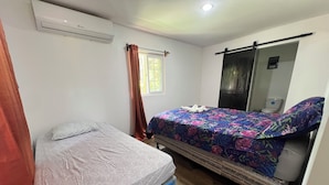 Habitación con dos camas, una con colchón y otra con colchón sobre ruedas. Hay una ventana con cortinas, un espejo y un equipo de aire acondicionado en la pared.