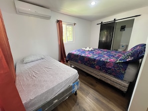 A room with two beds, one with a mattress and the other with a mattress on wheels. There is a window with curtains, a mirror, and an air conditioning unit on the wall. - Raul Villas  Hotel & Hostal  (Coxen Hole)