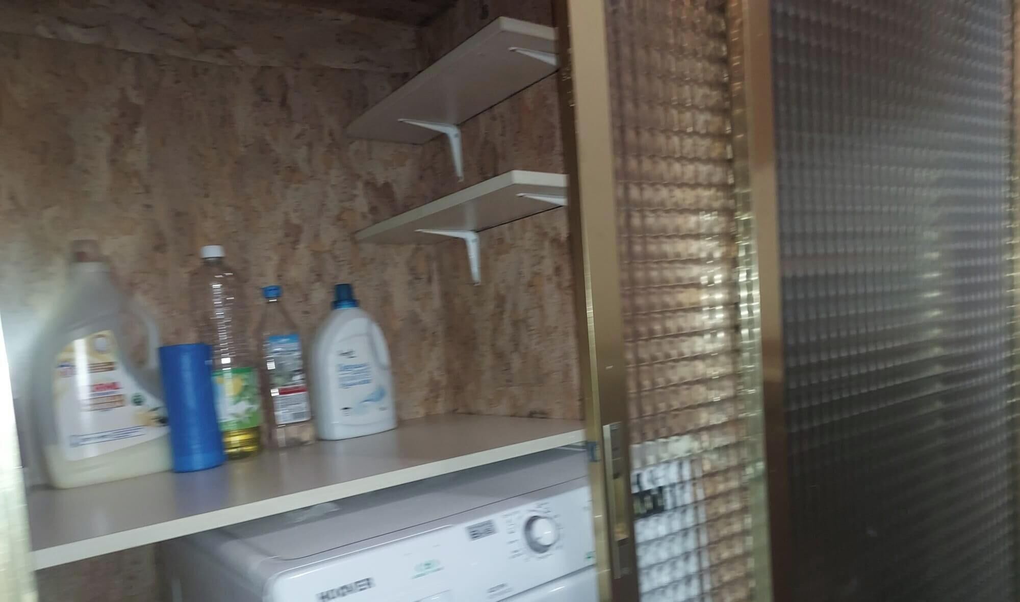 Een kamer met een wasmachine, planken met flessen en een metalen deur.