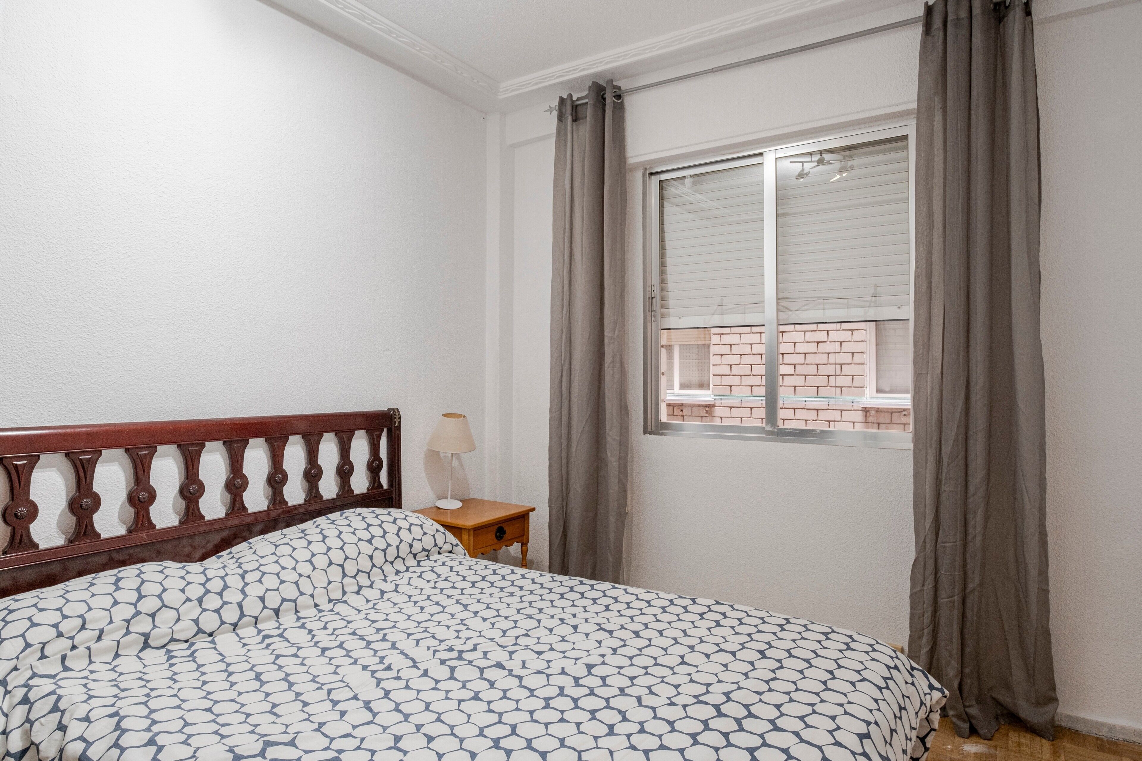 Un dormitorio con cabecera de madera, colcha estampada, mesita de noche con lámpara y ventana con cortinas.