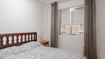 Un dormitorio con cabecera de madera, colcha estampada, mesita de noche con lámpara y ventana con cortinas.
