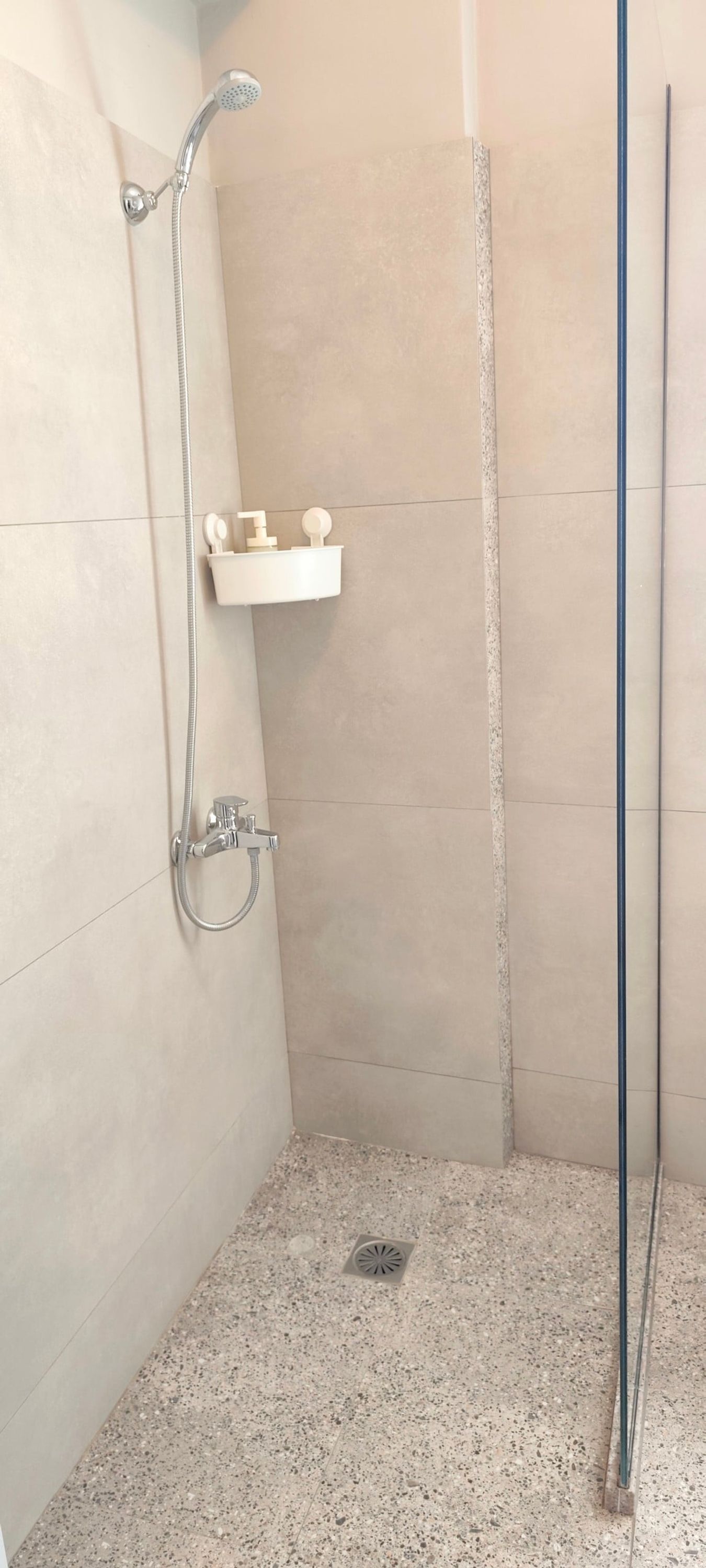Un petit espace douche fermé, doté d’un pommeau de douche à main, d’un robinet mural et d’une étagère pour les articles de toilette.