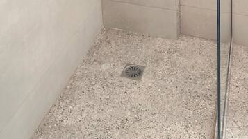 Un petit espace douche fermé, doté d’un pommeau de douche à main, d’un robinet mural et d’une étagère pour les articles de toilette.