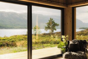 A room with large windows overlooking a lake and mountains, a wooden deck, and a cozy seating area with a plant.