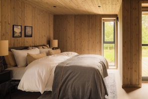 A bedroom with a wooden ceiling, a large bed with multiple pillows, a side table with a lamp, and a window with a view of greenery.